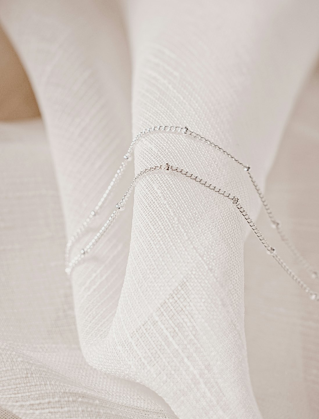 Two silver bead necklaces resting on white textured fabric.